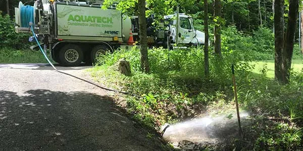 Aquatech sewer jetter cleans a roadside drainage area. Equipment parked on adjacent road, hose deployed in wooded setting.