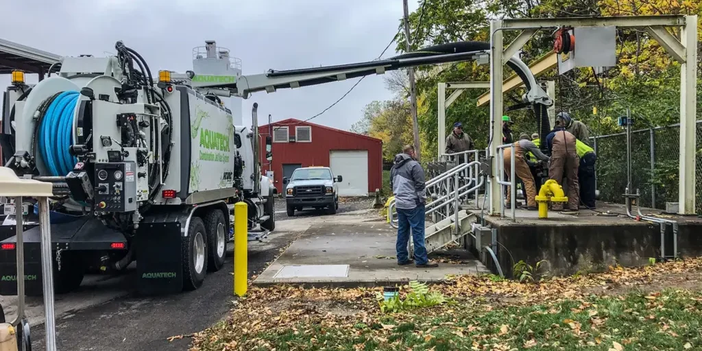 Aquatech combination sewer jetter in operation, with team conducting sewer cleaning work beside industrial facility and support truck.