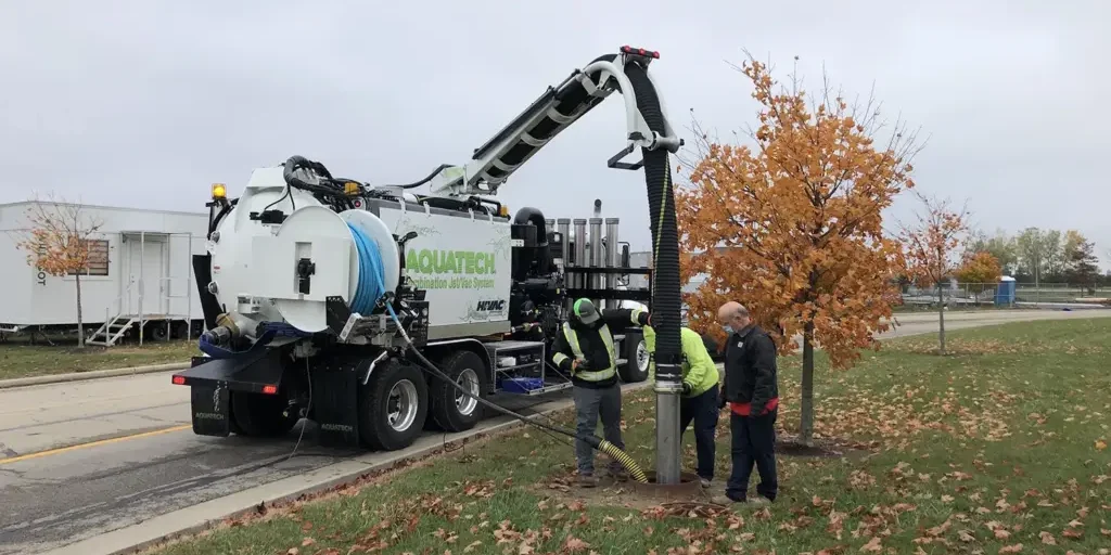 Aquatech combination sewer jetter truck operating, with operators vacuuming a manhole on a roadside, surrounded by autumn trees.