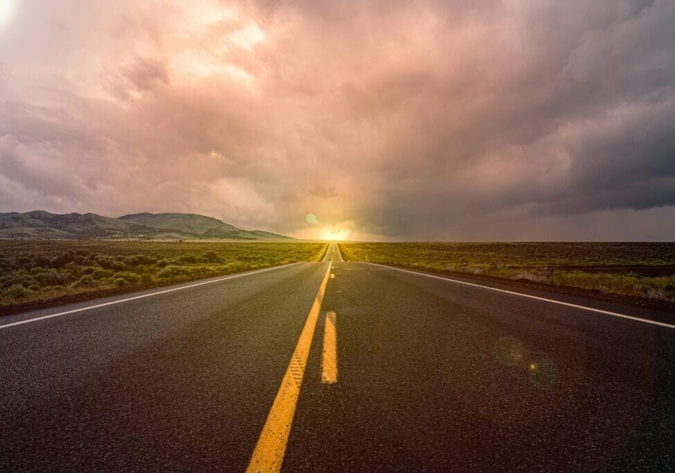 Open road stretching toward the horizon under a dramatic cloudy sky, with distant hills and evening sunlight in the background.