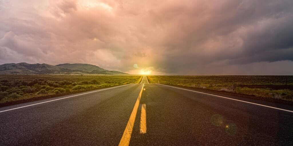 Open road stretching toward the horizon under a dramatic cloudy sky, with distant hills and evening sunlight in the background.